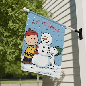 Może przedstawiać: Niebieska flaga zewnętrzna z zimową sceną z Charliem Brownem, Snoopy'm i bałwanem. Flaga ma czerwony napis "Let It Snow!". Postacie znajdują się w zaśnieżonym krajobrazie.