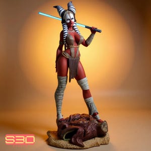 Puede incluir: Estatua detallada de una mujer alienígena de piel roja con marcas blancas y negras en la cabeza, sosteniendo un sable de luz azul. Viste un atuendo marrón y vendas blancas en las piernas, de pie sobre una base marrón detallada.