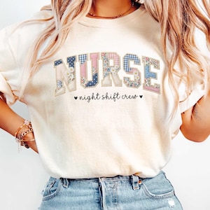 May include: Cream-colored sweatshirt with the word "NURSE" in patchwork letters. The letters feature floral, polka dot, and gingham patterns in blue, pink, and white. Below the word is the phrase "night shift crew" with heart symbols.
