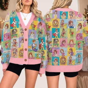 May include: A long-sleeve knit cardigan featuring a repeating grid pattern of Junie B. Jones illustrations in various colors. The garment has a V-neckline, a pink ribbed trim, and four brown buttons down the front. The text Junie B. Jones appears.