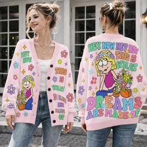 Puede incluir: Un cárdigan rosa claro con botones que presenta una ilustración de dibujos animados de un personaje rubio con una corona de flores sosteniendo un ramo. El texto dice HEY NOW HEY NOW THIS IS WHAT DREAMS ARE MADE OF, rodeado de flores y estrellas.