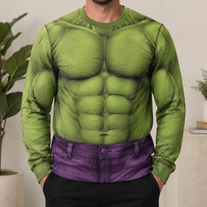 Puede incluir: Una sudadera de manga larga con cuello redondo que presenta un estampado gráfico de un torso verde musculoso con músculos abdominales y pectorales definidos, que se transforma en un diseño de cintura morada que imita un pantalón con botón.