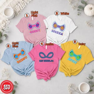 May include: Five short-sleeved t-shirts in light pink, white, light blue, azalea, and daisy yellow. Each shirt features a colorful mask graphic and a name: Ginny, Sparks, Buddy, Cat Burglar, or Bitsy. The shirts are arranged in a circular layout.