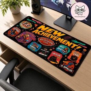 May include: A rectangular black desk mat featuring the large yellow text NEW ACHIEVEMENT! surrounded by various colorful graphic patches with illustrations and phrases including The Desperado Club, I am yelling Carl, and Mongo is appalled.