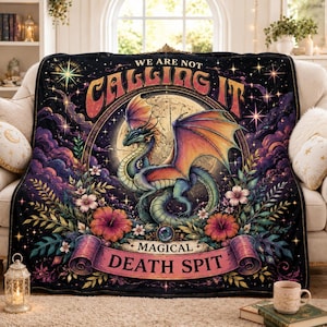 Op de afbeelding: Een zwarte plaid met een centrale illustratie van een draak met iriserende vleugels, omringd door een volle maan, paarse wolken en kleurrijke bloemen. De tekst luidt WE ARE NOT CALLING IT MAGICAL DEATH SPIT in gestileerde letters.