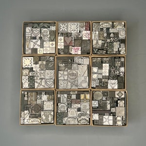 May include: Nine small square boxes filled with various black and white rubber stamps. The stamps are arranged in a grid pattern and feature different designs, including hearts, stars, and geometric shapes. The boxes are made of cardboard and have a brown color.