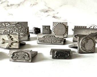 May include: A collection of vintage metal stamps featuring various designs, including a car, an airplane, a skull and crossbones, a gear, a globe, and a stylized letter 'S'.