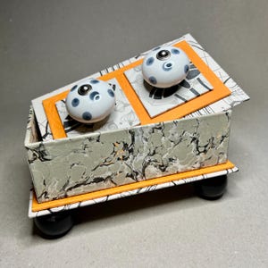 May include: A decorative box with a hinged lid, featuring a marbled gray and black pattern on the sides. The lid has two white and blue polka-dot knobs and is accented with orange and patterned paper. The box rests on four black spherical feet.