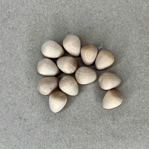 May include: A group of 12 smooth, light brown wooden eggs. The eggs are arranged in a loose circle on a gray textured surface.