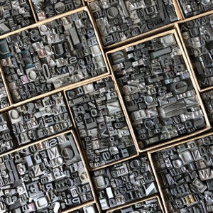May include: A close-up view of a collection of vintage metal letterpress printing blocks in various sizes and styles. The blocks are arranged in a grid pattern and are contained in small wooden boxes.