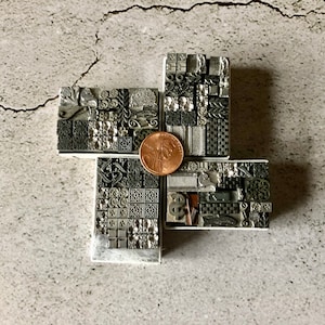 May include: A set of small, square, metal printing blocks with various designs, arranged in a cross shape. The blocks are in shades of gray and black, and a copper-colored coin is placed in the center of the cross.