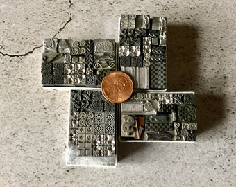 May include: A set of small, square, metal printing blocks with various designs, arranged in a cross shape. The blocks are in shades of gray and black, and a copper-colored coin is placed in the center of the cross.