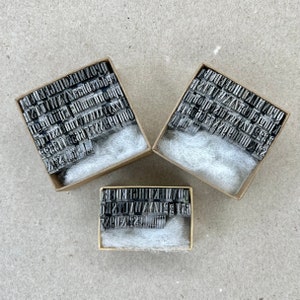 May include: Three boxes filled with metal letter and number stamps. The boxes are made of cardboard and have a white, fluffy material inside. The stamps are arranged in alphabetical and numerical order.
