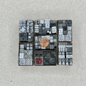 May include: A collection of 36 small, square, metal printing blocks with various geometric and floral designs. The blocks are arranged in a grid pattern and are in shades of gray and black. A single copper-colored coin is placed in the center of the grid.