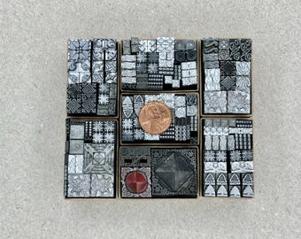May include: A collection of 36 small, square, metal printing blocks with various geometric and floral designs. The blocks are arranged in a grid pattern and are in shades of gray and black. A single copper-colored coin is placed in the center of the grid.