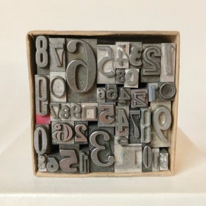 May include: A collection of vintage metal letterpress printing blocks, including numbers and symbols, neatly arranged inside a cardboard box. The blocks are various sizes and fonts, with a worn, industrial aesthetic. The box is a light brown color.