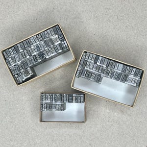 May include: Three open cardboard boxes filled with individual metal letterpress type. The type is arranged in rows, showing various letters, numbers, and punctuation marks. The boxes are on a light gray surface.