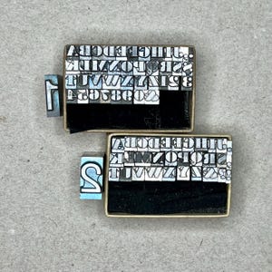 May include: Two vintage metal letterpress trays filled with individual metal letter blocks. The trays are rectangular and made of cardboard. The letter blocks are silver and black. Two individual letter blocks are visible outside the trays.