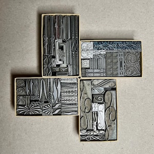May include: Four rectangular cardboard boxes filled with an assortment of metal letterpress printing blocks. The blocks feature various engraved patterns, including floral motifs, geometric lines, scrollwork, and decorative borders in silver tones.