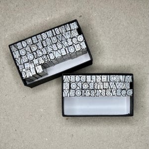 May include: Two open black boxes filled with metal letterpress type. The type is arranged in rows and includes letters, numbers, and symbols. The boxes are on a light gray surface.