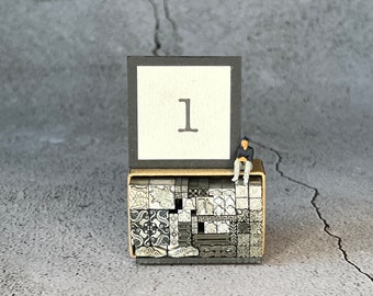 May include: A black frame with a white background and the number "1" printed in black. The frame is sitting on top of a stack of small, square, metal blocks with intricate designs. A miniature figurine of a person is sitting on top of the frame.
