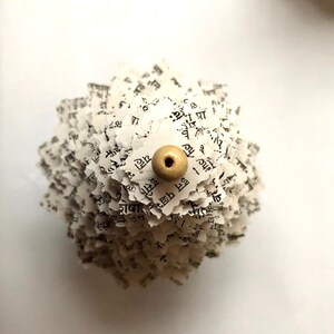 May include: A white paper flower made from pages of a book with a beige bead in the centre.