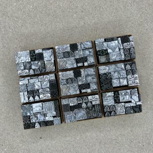 May include: A set of nine small boxes filled with various metal printing blocks. The blocks are arranged in a grid pattern and feature a variety of designs, including floral motifs, geometric shapes, and abstract patterns. The blocks are all silver in color and have a slightly textured surface.
