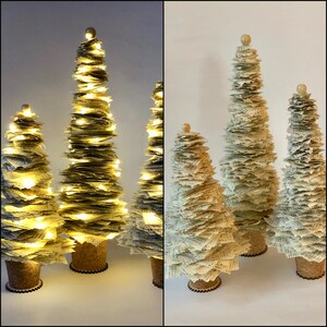 May include: Three paper Christmas trees decorated with white paper and topped with a small wooden ball. The largest tree is lit with white fairy lights.