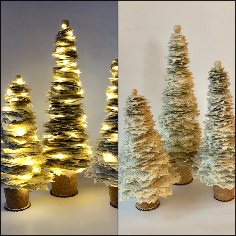 May include: Three paper Christmas trees decorated with white paper and topped with a small wooden ball. The largest tree is lit with white fairy lights.