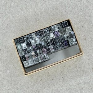 May include: A wooden box filled with metal letterpress printing blocks. The blocks are arranged in rows and columns, and they contain the letters of the alphabet, numbers, and punctuation marks.
