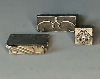 May include: Three vintage metal printing blocks with intricate designs. The blocks are made of a dark metal and have a silver-colored design. The designs include a swirl pattern, a geometric pattern, and a floral pattern.