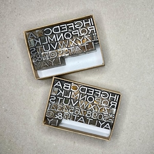 May include: Two open cardboard boxes filled with metal letterpress type. The letters and numbers are arranged in rows. The top box has the word "YAY" visible. The type is silver-colored and the boxes are beige.
