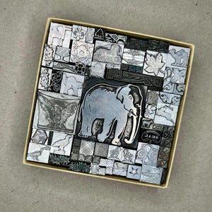 May include: A collection of vintage metal printing blocks in a cardboard box. The blocks feature various designs, including an elephant, an eagle, and other geometric and animal motifs. The blocks are silver-colored and arranged in a grid pattern.
