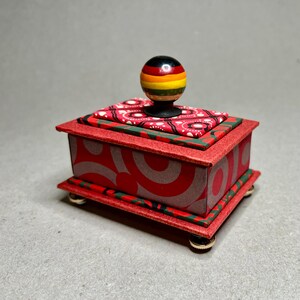 May include: A small, red decorative box with a patterned design. The box has a lid with a colourful, striped knob. The sides of the box feature a red and grey swirl pattern. The box sits on small, round feet.