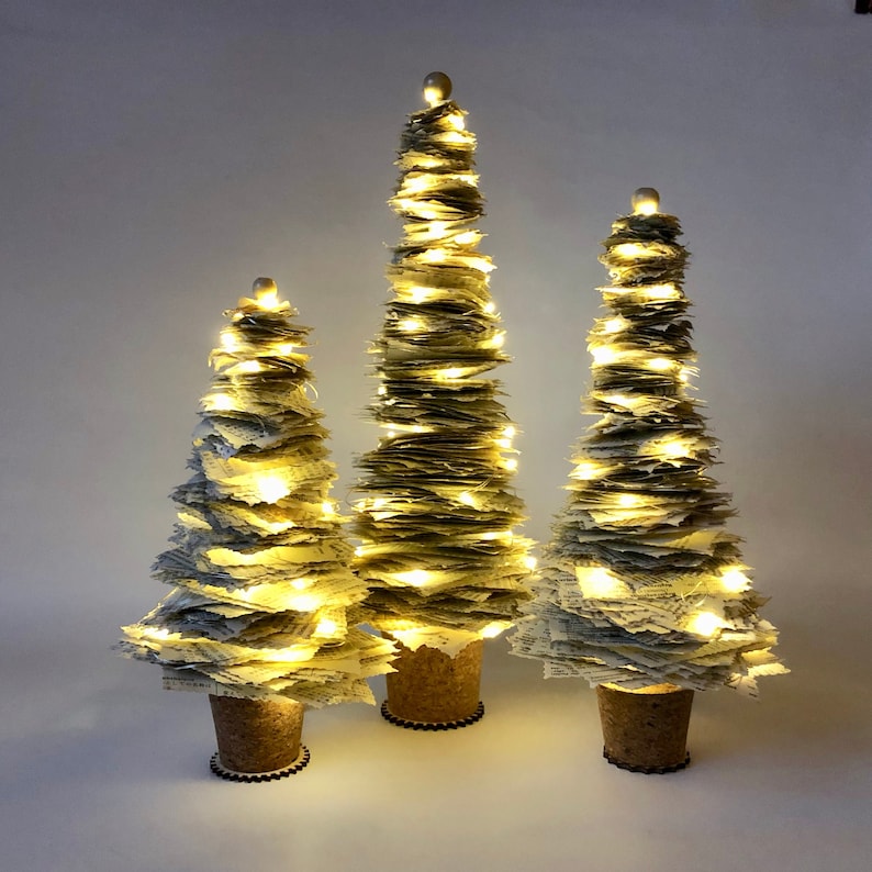 May include: Three white paper Christmas trees with warm white lights. The trees are different sizes and are arranged in a row on a white background.
