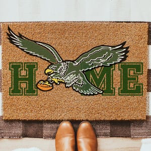 May include: A doormat with a brown background and the word "HOME" in green block letters. A green and white eagle graphic is centered above the word "ME". The mat is on a black and white checkered rug. Brown boots are visible.