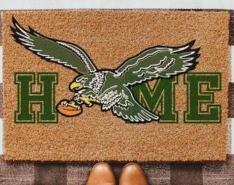 May include: A doormat with a brown background and the word "HOME" in green block letters. A green and white eagle graphic is centered above the word "ME". The mat is on a black and white checkered rug. Brown boots are visible.