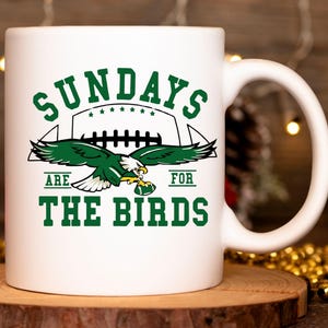 Op de afbeelding: Witte keramische mok met een groene en witte afbeelding. De afbeelding toont een adelaar met een voetbal en de tekst "SUNDAYS ARE FOR THE BIRDS". De mok staat op een houten onderzetter.