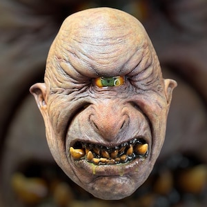 May include: A grotesque, one-eyed mask with a menacing expression. The mask features a wrinkled, tan complexion, a single, yellow-green eye, and a snarling mouth with decaying teeth. Drool is visible. The overall theme is horror or fantasy.