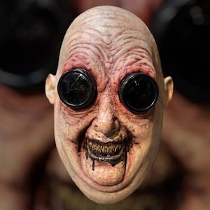 May include: A grotesque, bald mask with large, black, circular lenses over the eyes. The mask has a wrinkled, flesh-toned appearance with red accents around the eyes and mouth. The mouth is open, revealing sharp, yellow teeth and a dark, dripping substance.