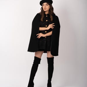 Puede incluir: Una capa negra con botones dorados y una boina a juego. La capa se lleva sobre un vestido negro y se combina con botas altas negras. El conjunto se presenta sobre un fondo blanco, creando un look clásico y elegante.