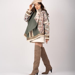 Op de afbeelding: Een vrouw draagt een beige baret, een jas met patroon met een groene drape en kniehoge bruine suède laarzen. De jas heeft kleurrijke bloemenborduursels en franjes. De laarzen hebben een spitse neus en een kleine hak.
