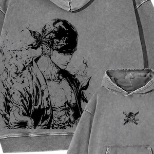 May include: A charcoal gray acid-wash hoodie featuring a large black ink-style illustration of a character wearing a bandana on the back and a small skull and crossbones emblem on the front chest, complete with a hood and a front kangaroo pocket.