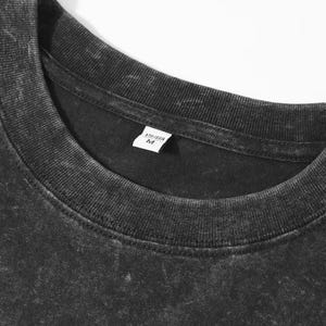 May include: Close-up of a black, textured t-shirt with a crew neck. A small white tag near the collar displays the size and other information. The shirt has a faded, vintage appearance.