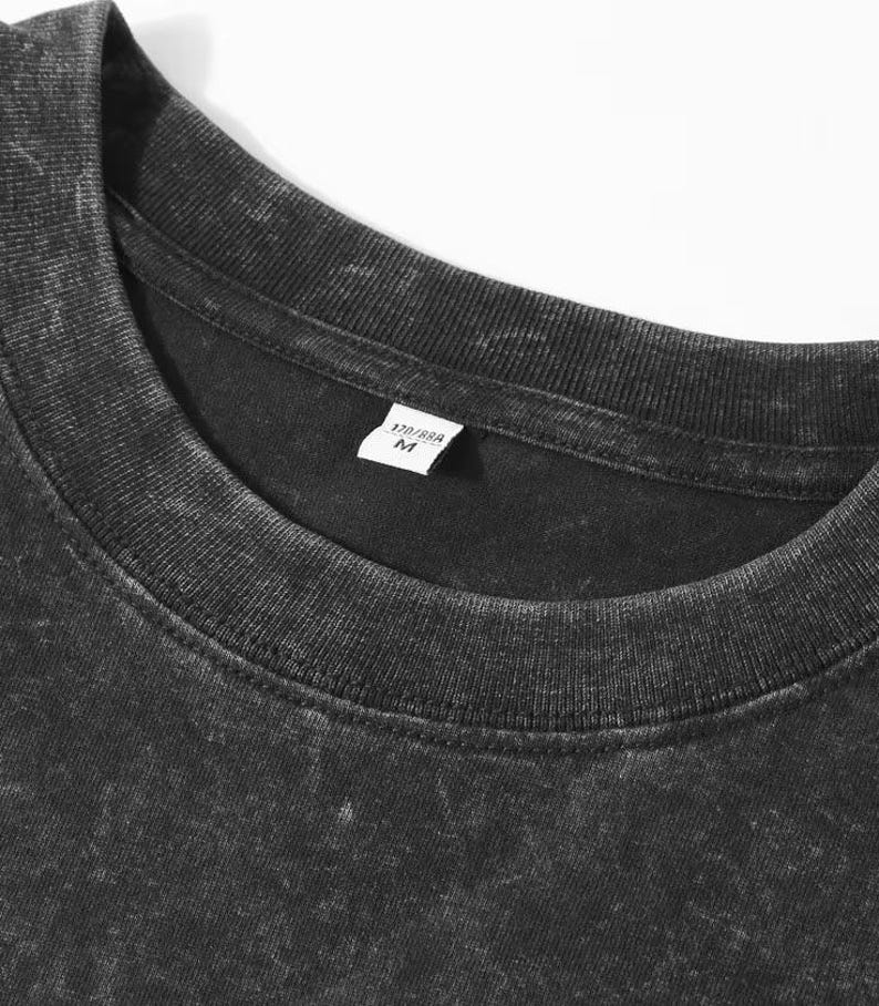 May include: Close-up of a black, textured t-shirt with a crew neck. A small white tag near the collar displays the size and other information. The shirt has a faded, vintage appearance.