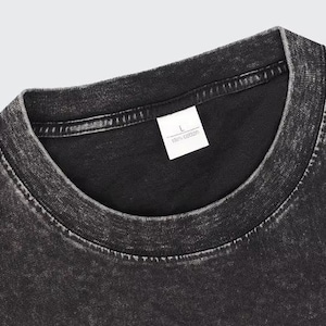 May include: Close-up of a black, acid-wash style t-shirt. The collar and shoulder area are visible, with a white tag that reads "L" and "100% cotton". The fabric has a textured, worn appearance.