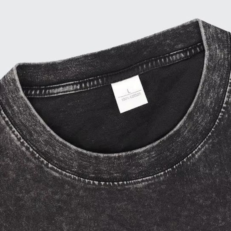 May include: Close-up of a black, acid-wash style t-shirt. The collar and shoulder area are visible, with a white tag that reads "L" and "100% cotton". The fabric has a textured, worn appearance.