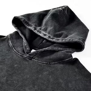 May include: Close-up of a dark gray hoodie with a distressed, vintage wash effect. The hood is up, revealing the interior lining. The fabric has a textured appearance, and the overall style suggests casual wear.