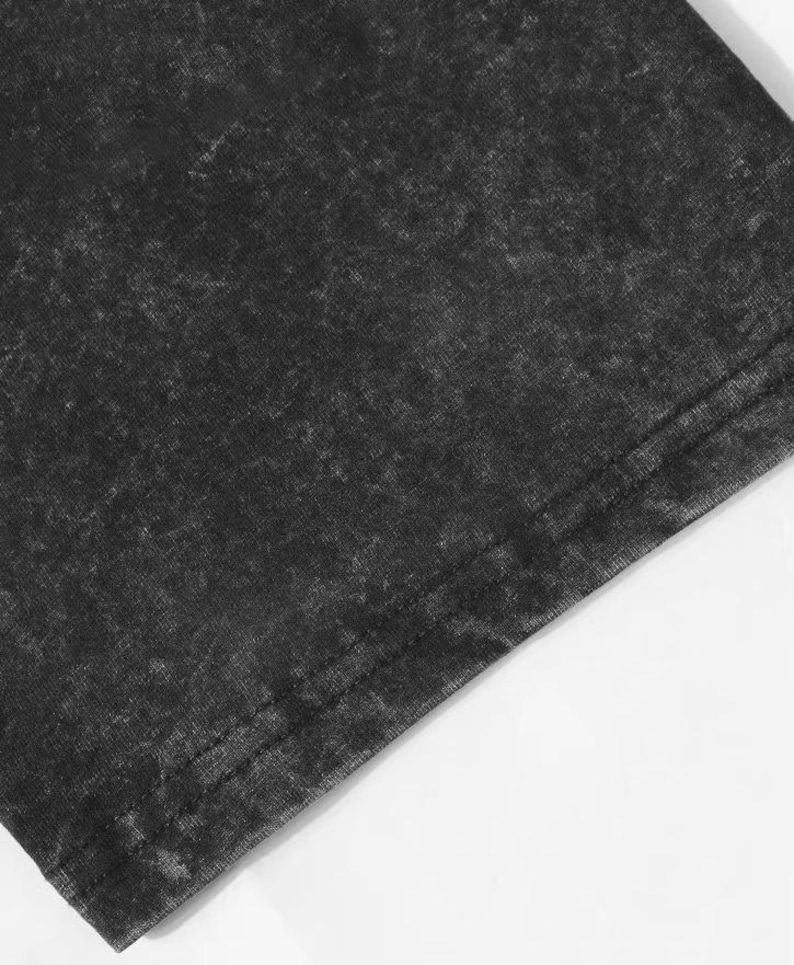 May include: Close-up of a section of dark grey, acid-washed fabric. The fabric has a textured appearance with lighter grey areas, and a visible seam along the edge. The material appears to be a t-shirt or similar garment.