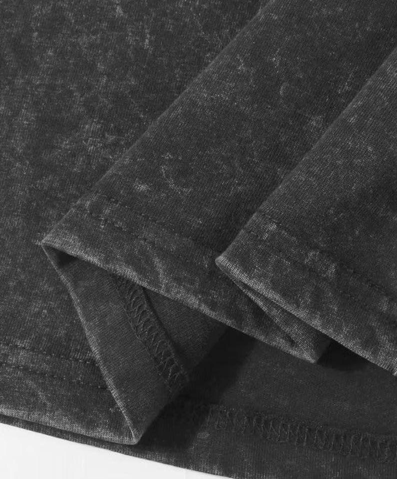 May include: Close-up of a dark grey, acid-wash style t-shirt. The fabric is folded, showing the hem and seams. The texture of the material is visible, with a slightly faded appearance. The shirt appears to be made of a soft, comfortable fabric.